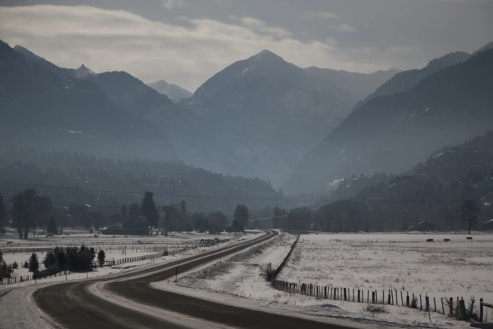 A winding road crosses snow-covered fields toward hazy mountains.